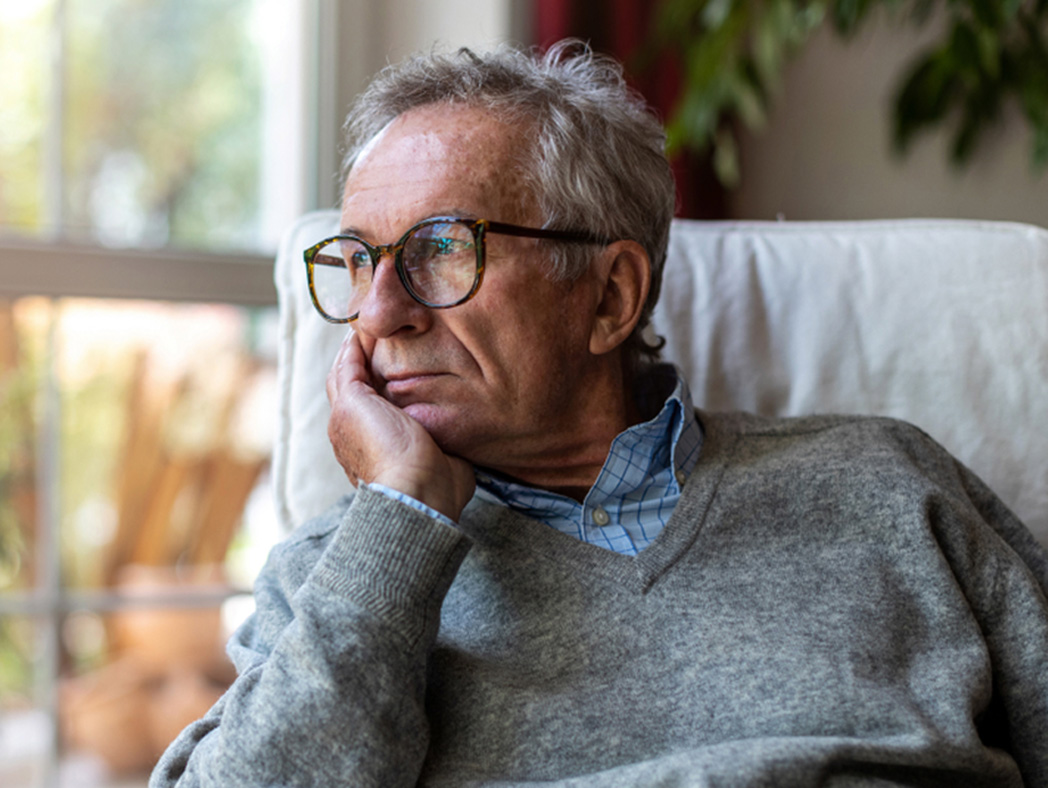 Older man sitting down and looking out window, holding his face in one hand.