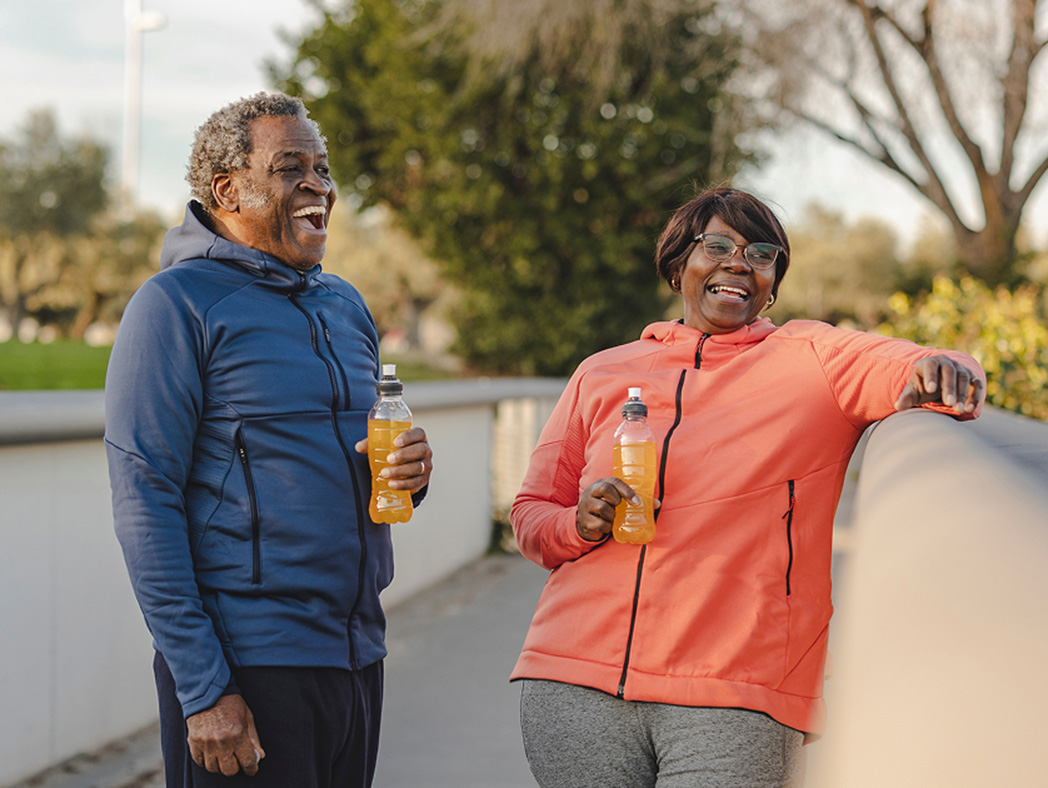An older couple standing together outside laughing and drinking an orange beverage from a plastic water bottle.