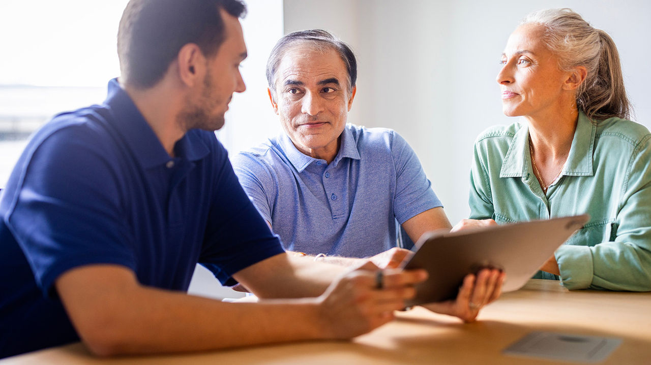 Elderly couple sitting down and conversing with a middle-aged man holding a digital tablet device.